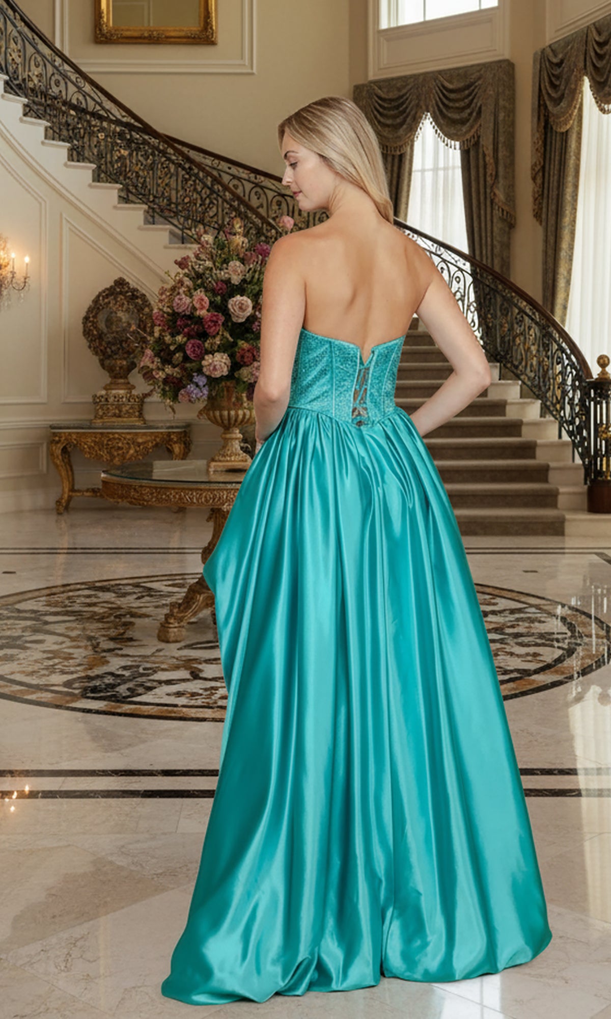 A woman stands indoors, facing away, wearing the Long Prom Dress: Poly USA 25156—a strapless turquoise satin gown with a rhinestone bodice and full skirt—in a grand room with a staircase and floral arrangement.